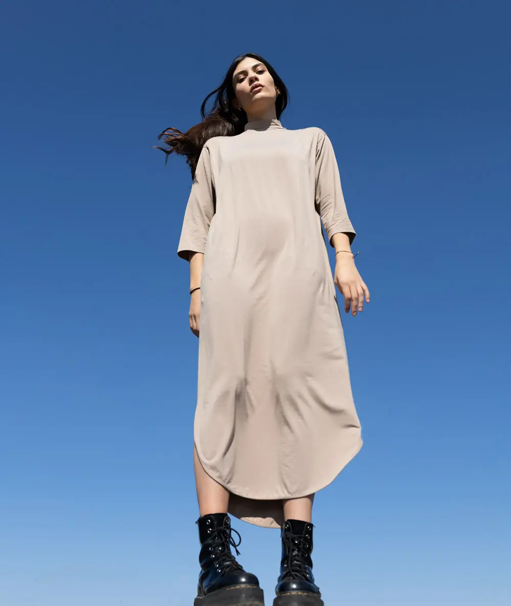 
                   Model in beigem Kleid und schwarzen Stiefeln steht unter klarem blauem Himmel.
                   